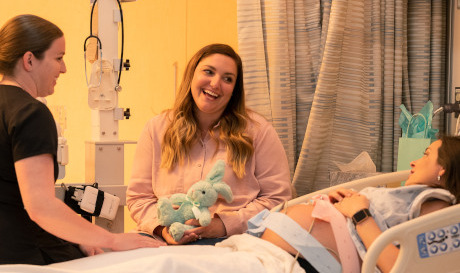 Dos mujeres sentadas con una mujer embarazada en el hospital, todas sonriendo y hablando juntas.